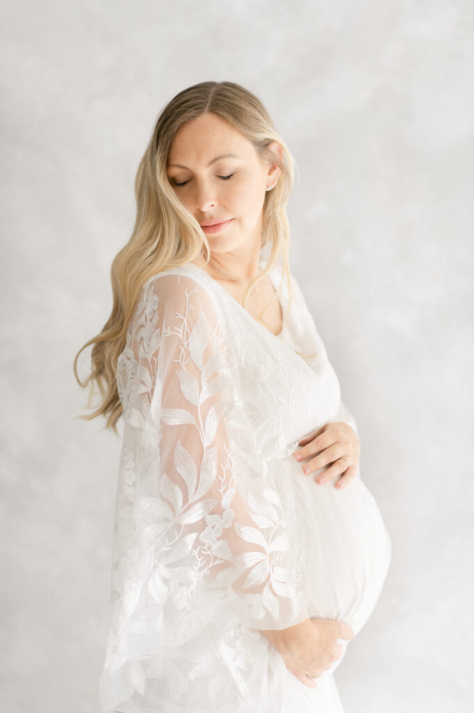Image of pregnant mother with long blonde hair wearing a beautiful lace white dress, holding her pregnant belly and looking down softly, as photographed by Sydney's best maternity photographer Sarah Vassallo Photography