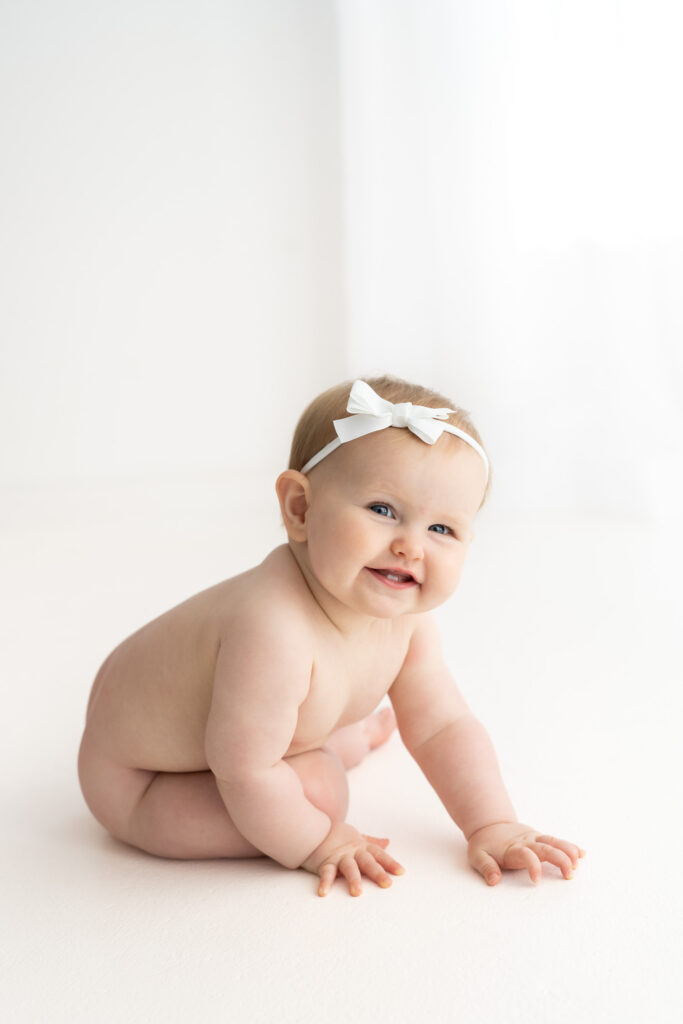 Image of little baby girl with a white bow, sitting in a white studio grinning cutely at camera as photographed by Sydney's best baby photographer Sarah Vassallo