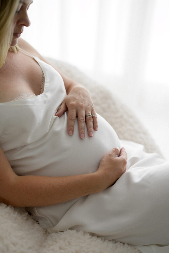 Closeup image of pregnant woman sitting in white silk dress holding her belly as photographed by Sutherland Shire maternity photographer Sarah Vassallo