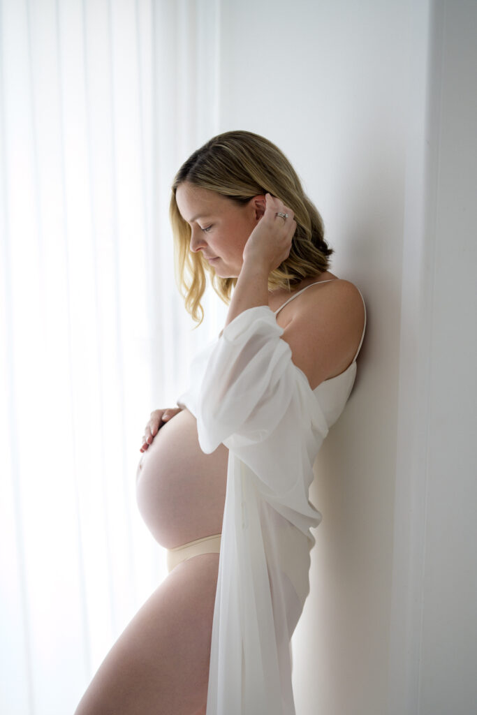 Pregnant mother looking down at her belly while leaning on wall in white studio in Sutherland Shire Sydney