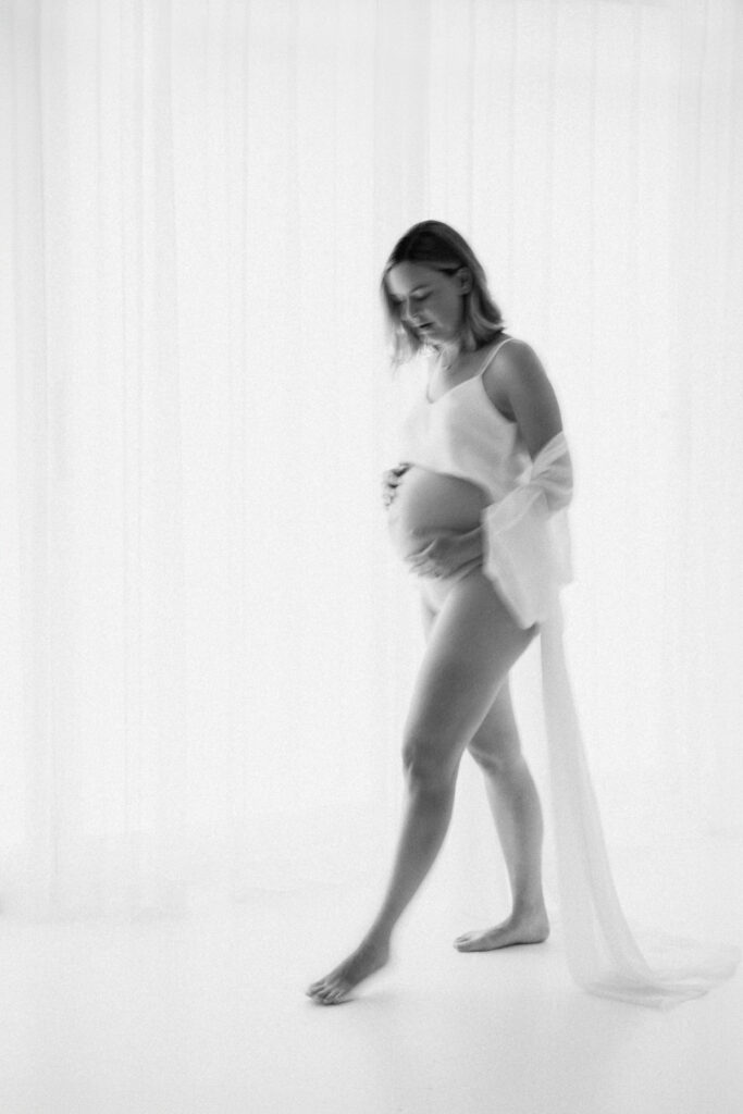 black and white image of pregnant mother walking in white studio holding her belly gently, with artistic blur to the image, as photographed by Sydney maternity photographer Sarah Vassallo
