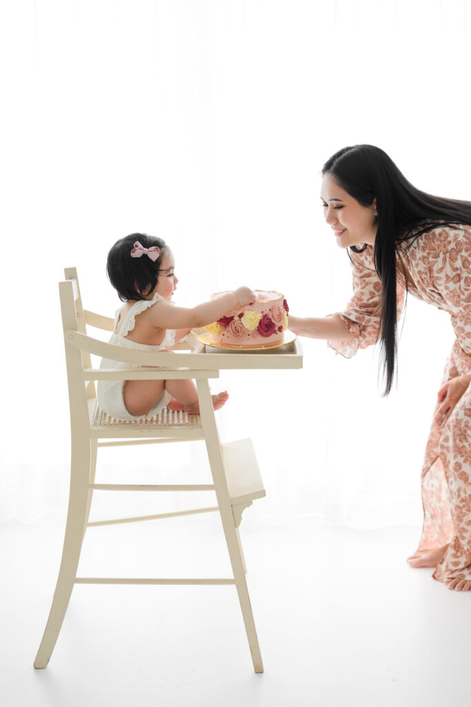 Baby sitting in highchair while her mum hands her a first birthday cake during a milestone session in the Sutherland Shire