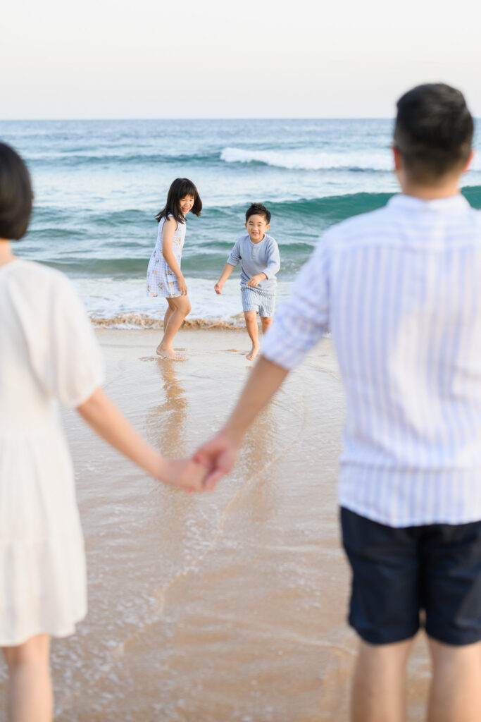 Parents holding hands on the beach, and kids playing in the water beyond them during family photo session in Sydney
