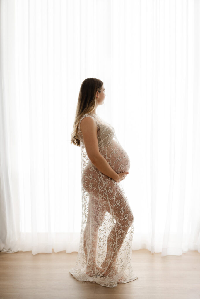 Backlit maternity portrait of young mother wearing sheer dress, gazing out the window during maternity photo session in Sydney
