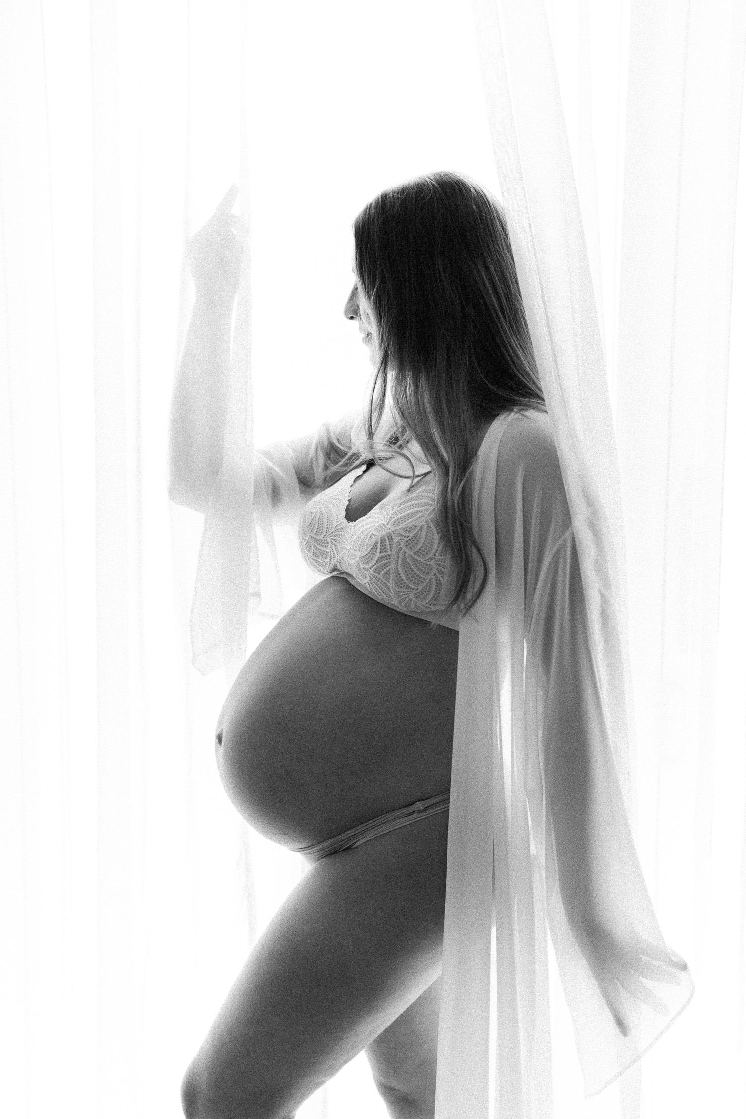 Elegant black and white photo of pregnant mother standing by sheer curtains, belly visible in robe as photographed by Sydney maternity photographer Sarah Vassallo