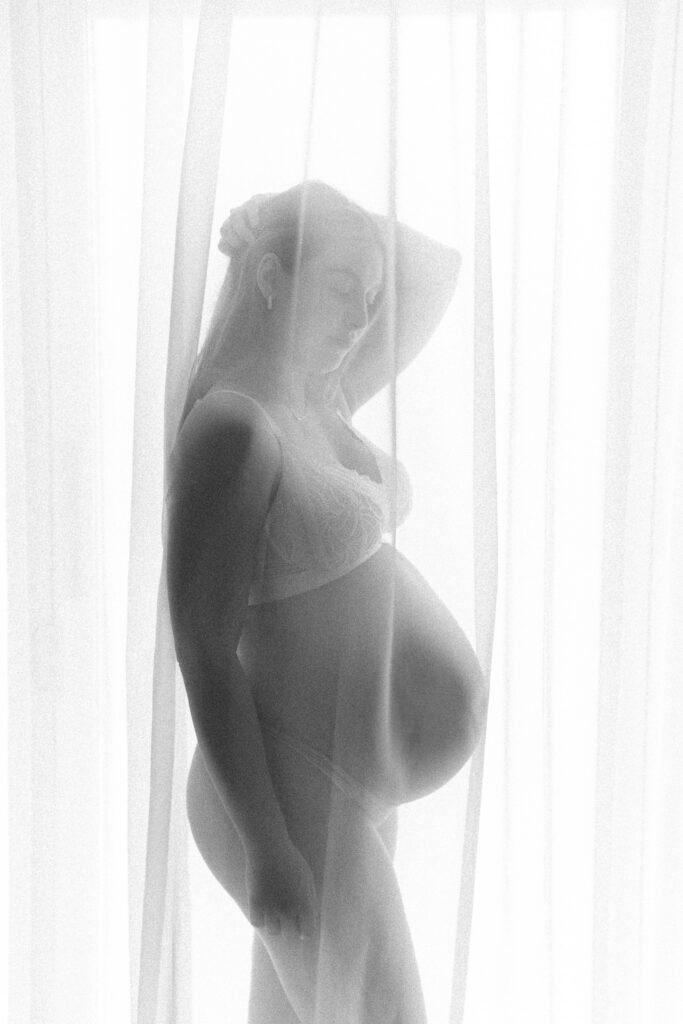 Elegant black and white photo of pregnant mother standing behind sheer curtains, belly visible