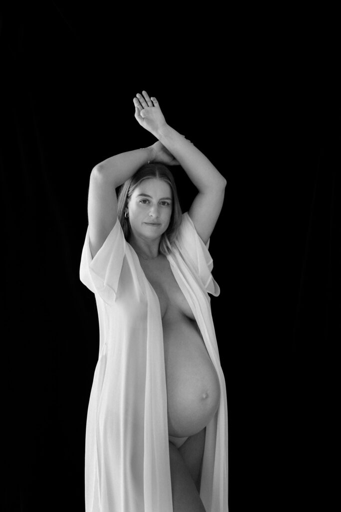Pregnant mother wearing white robe, posed against black backdrop in black and white image photographed in Sutherland Shire maternity studio